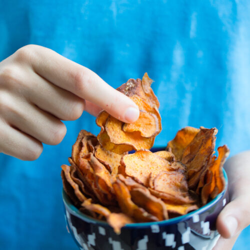 A person wearing a blue shirt holds a patterned bowl filled with thin, crispy, healthy sweet potato chips. The left hand supports the bowl as the right hand picks up a chip. The softly blurred background keeps the focus on the snack.
