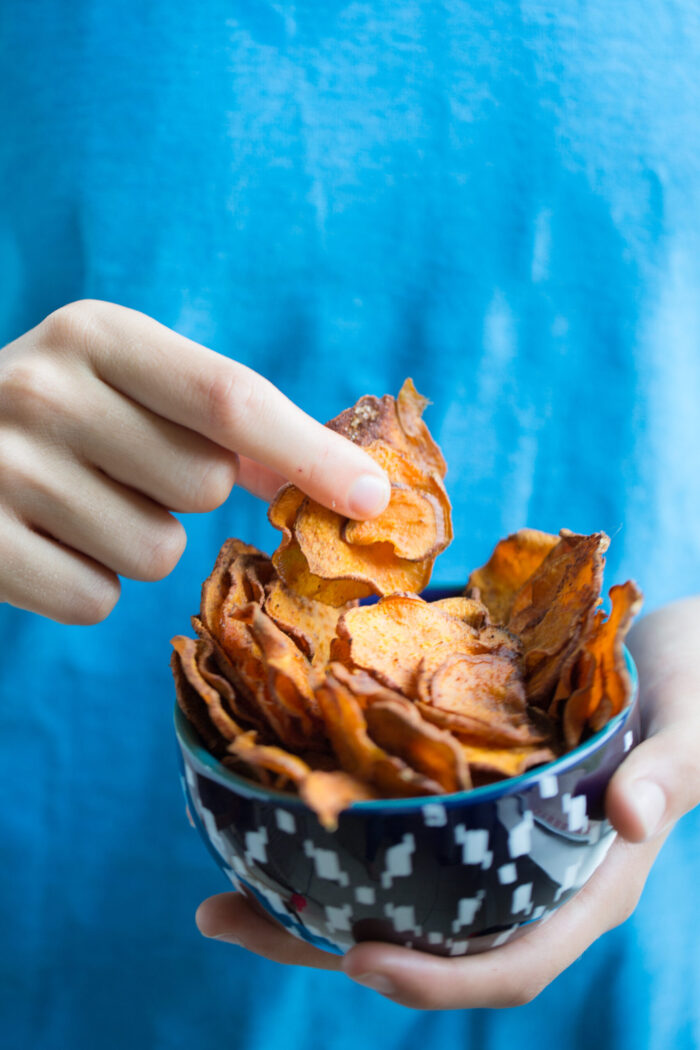A person wearing a blue shirt holds a patterned bowl filled with thin, crispy, healthy sweet potato chips. The left hand supports the bowl as the right hand picks up a chip. The softly blurred background keeps the focus on the snack.