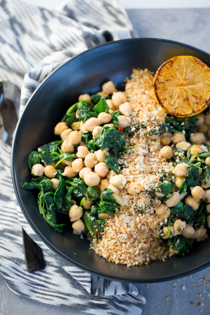 Chickpeas with spinach in a black bowl topped with bread crumbs and a yellow lemon on the side