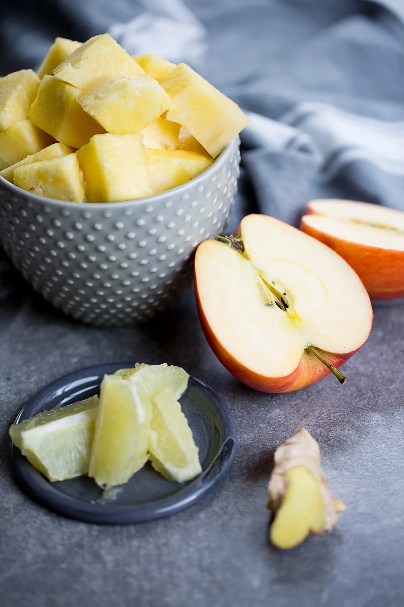A textured gray bowl filled with pineapple chunks is placed on a gray surface. Beside it, a halved apple and apple slices rest nearby. A small gray plate holds additional pineapple pieces for a perfect pineapple ginger juice blend, and a piece of ginger is also present. A soft focus striped cloth is in the background.