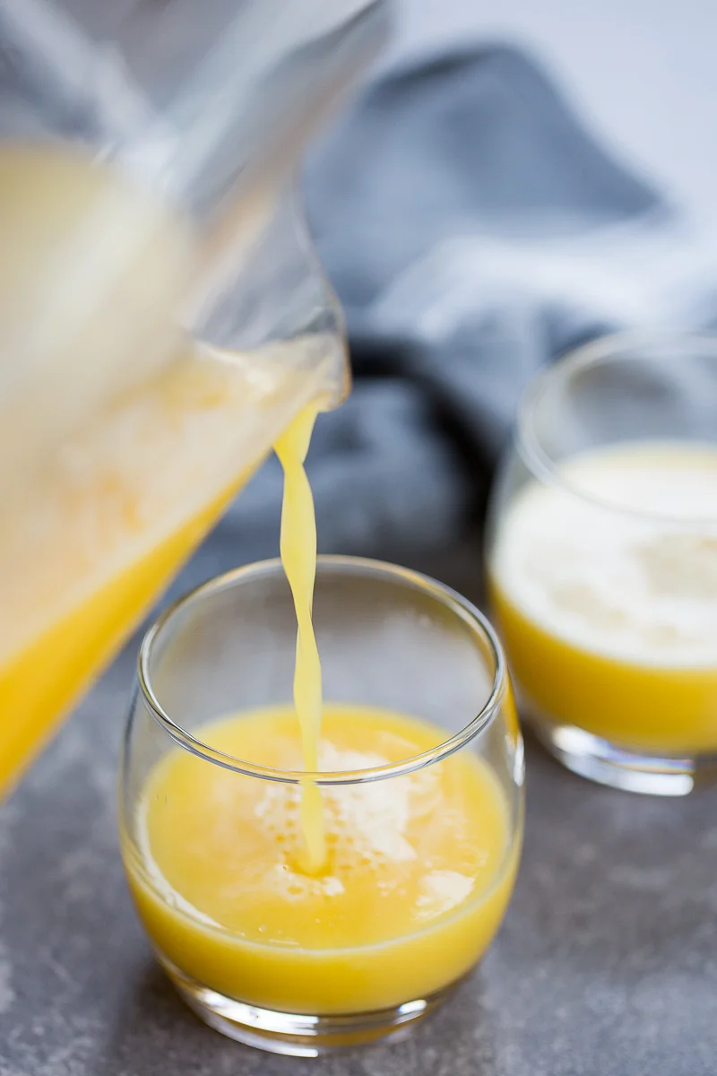Close-up of a pitcher pouring pineapple ginger juice into one of two clear round glasses. The glasses sit on a gray surface, with a soft-focus blue-gray cloth in the background. The juice's bright orange-yellow hue creates a refreshing and inviting appearance.