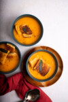 Three bowls of butternut squash soup with croutons, garnished with a sage leaf, presented on a light background with a red napkin and a spoon at the side.