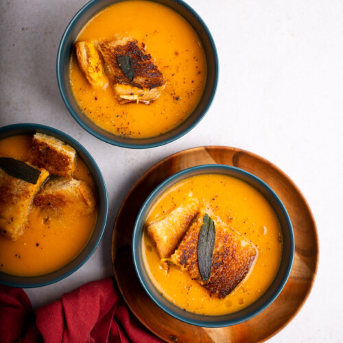 Three bowls of butternut squash soup with croutons, garnished with a sage leaf, presented on a light background with a red napkin and a spoon at the side.