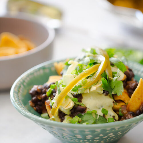 A green ceramic bowl filled with nachos, topped with black beans, poblano cashew crema, sliced yellow peppers, and fresh cilantro. The vibrant colors and textures are captivating, reminiscent of a healthy vegan Frito pie, with another bowl of chips blurred in the background.