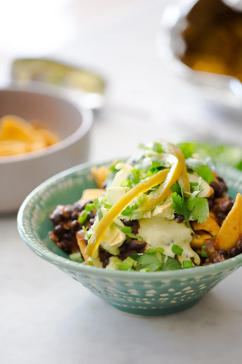 A green ceramic bowl filled with nachos, topped with black beans, poblano cashew crema, sliced yellow peppers, and fresh cilantro. The vibrant colors and textures are captivating, reminiscent of a healthy vegan Frito pie, with another bowl of chips blurred in the background.
