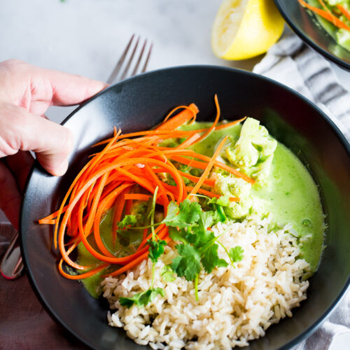 A black bowl with white rice, green Thai curry with tofu, and julienned carrots, garnished with fresh cilantro. A hand holds the bowl over a light cloth napkin, with a lemon wedge and another bowl partly visible in the background.