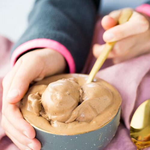 Una persona sostiene un cuenco gris lleno de cremoso helado de chocolate vegano y sirve un poco con una cuchara dorada. La persona lleva una manga oscura con un puño rosa, y una servilleta malva doblada descansa debajo del bol. Otra cuchara dorada descansa cerca, sobre una superficie blanca.