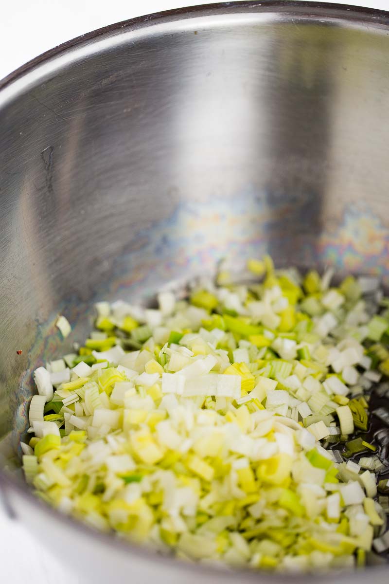 A close-up of a metal pot containing finely chopped leeks, ready for Red Lentil Soup. The pale green and white leeks glisten in the slightly rainbow-tinted pot, suggesting the start of comforting soup preparation.