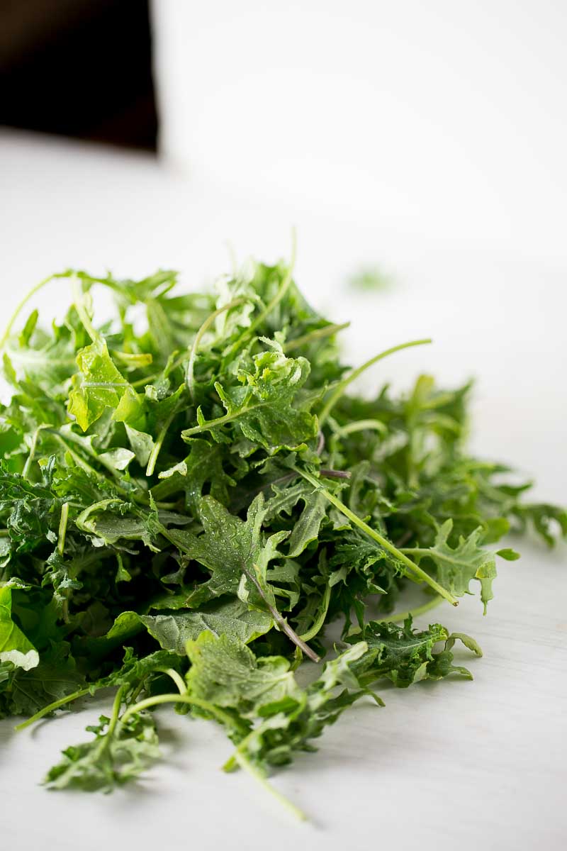 A close-up of a pile of fresh, leafy mixed greens, including arugula and kale, on a white surface. The leaves are vibrant green with varied textures and edges, reminiscent of ingredients perfect for a hearty Lentil Soup.