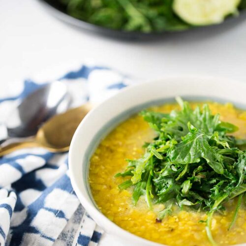 A white bowl filled with yellow lentil soup, inspired by a Red Lentil Soup Recipe, is topped with fresh arugula and sits on a white surface. Beside it are a blue-and-white checkered napkin and spoon; in the background, more arugula and a halved lemon.