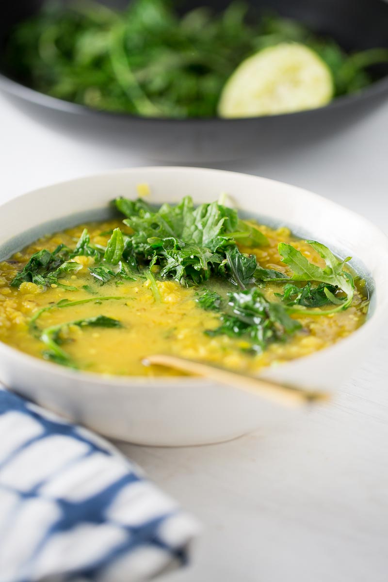 A bowl of yellow lentil soup topped with fresh leafy greens, possibly sautéed kale, sits on a white surface. A spoon rests in the bowl, beside a striped blue-and-white napkin. A blurred pan with greens and lemon slice is in the background.