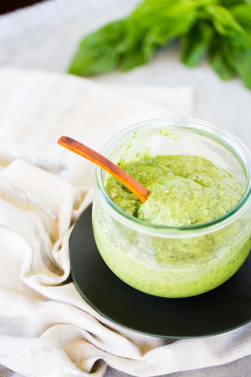 Vegan avocado pesto in a jar over a dark green saucer with fresh basil on the back.