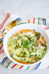 A white bowl of soup garnished with avocado slices, lime wedges, spiralized zucchini, chopped cilantro, and shredded greens sits on a striped cloth with a gold spoon beside it—perfect inspiration for your next chia recipe.