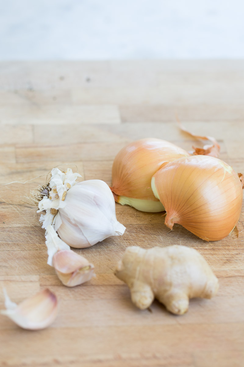 A whole head of garlic with separated cloves, a partially peeled ginger root, and two yellow onions with papery skin rest on a light wooden surface—fresh ingredients perfect for making a vibrant red curry. The background is softly blurred to highlight the ingredients.