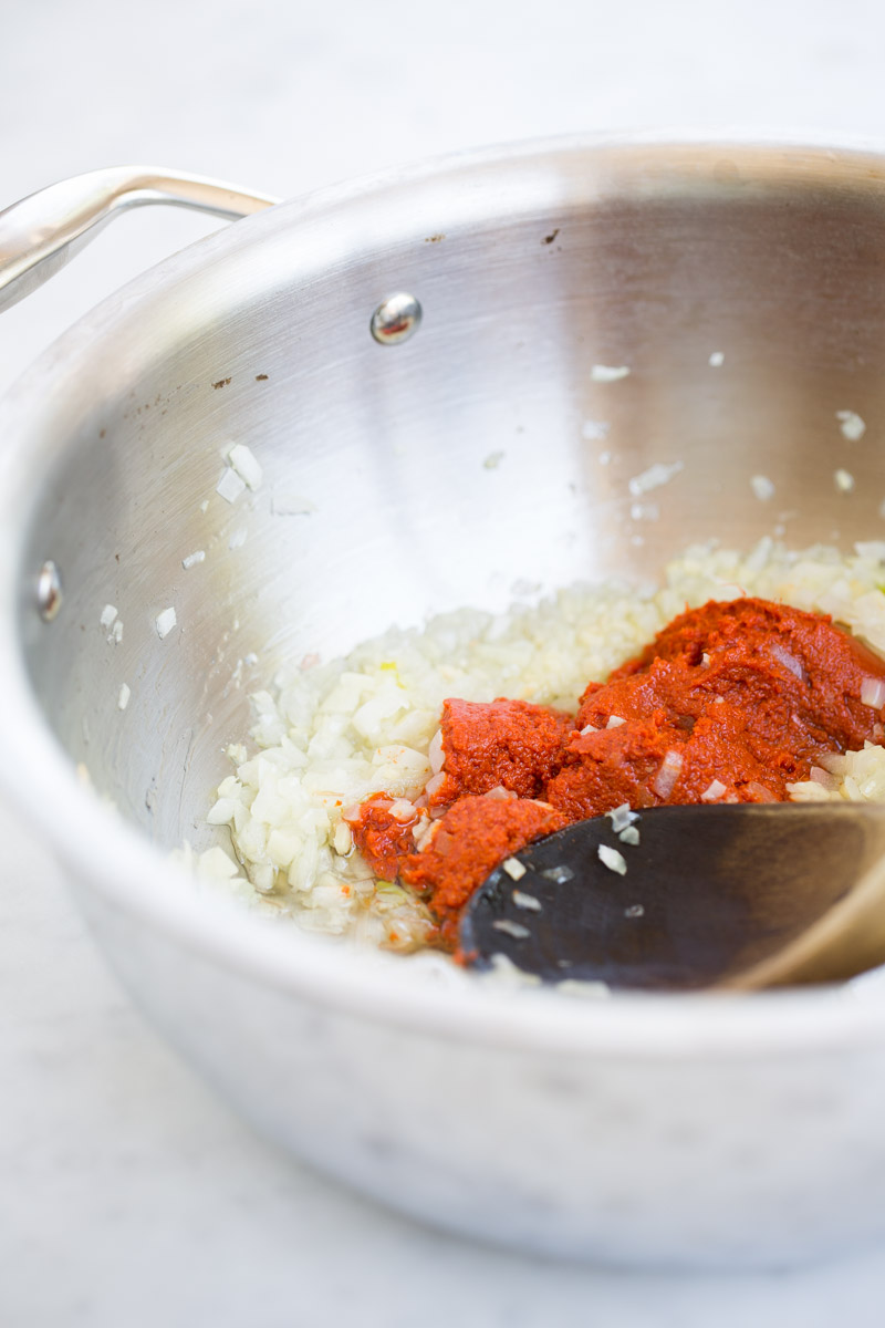 A stainless steel saucepan holds sautéing chopped onions with a spoonful of red curry paste added on top. A wooden spoon rests inside the pan, partially covered by the onions and curry paste. The background is a smooth, light surface.