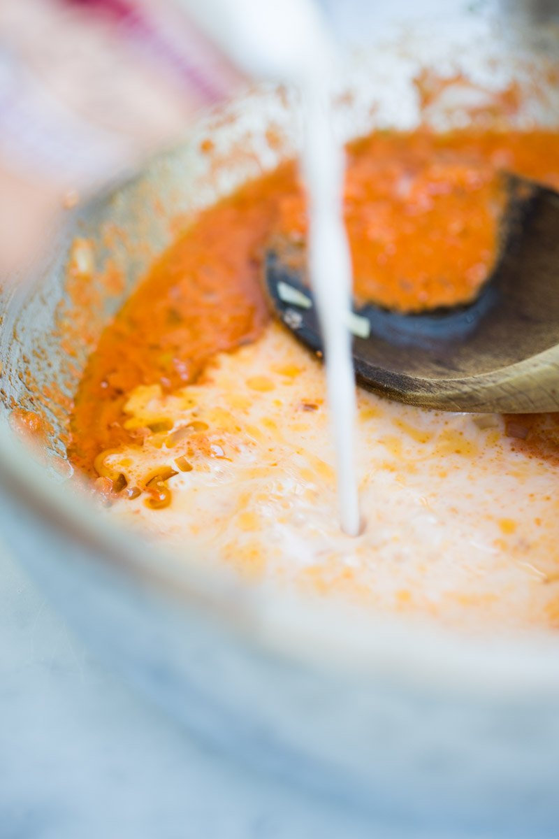 Close-up of a wooden spoon stirring a creamy liquid into a thick orange-red curry sauce in a glass bowl. The creamy swirl creates a beautiful marbled effect over the vibrant curry, while the softly blurred background adds warmth.