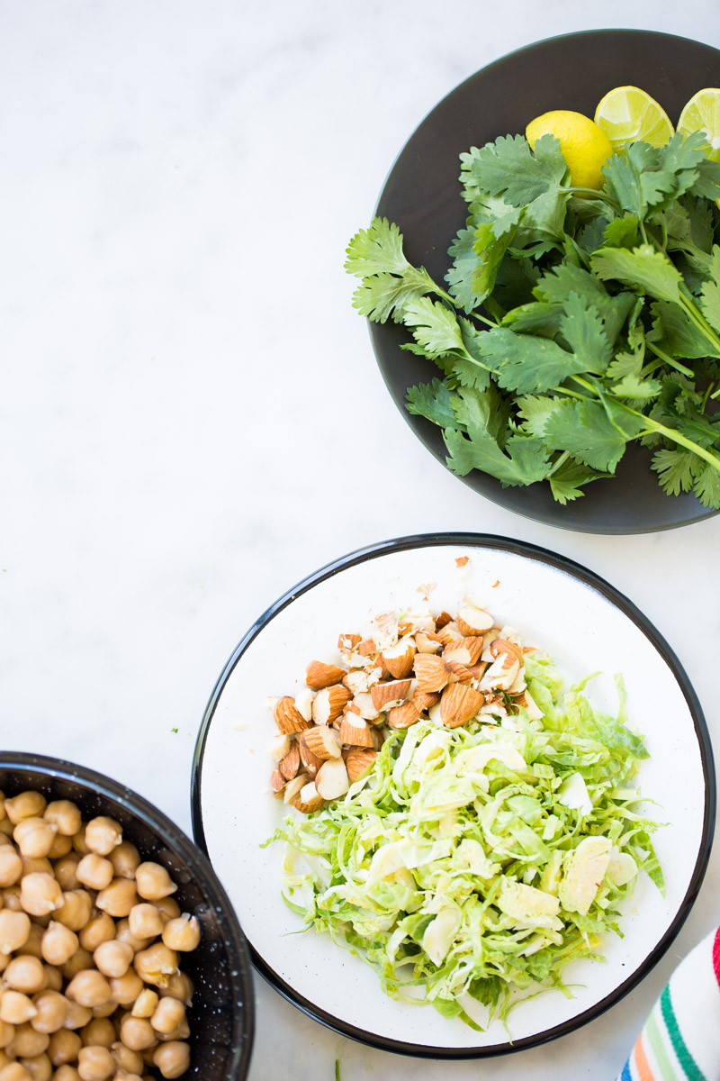 A white plate holds shredded Brussels sprouts and chopped almonds. Nearby, a black bowl is filled with chickpeas, ready for the red curry. At the top right, a black plate contains fresh cilantro and lemon slices on a white surface.