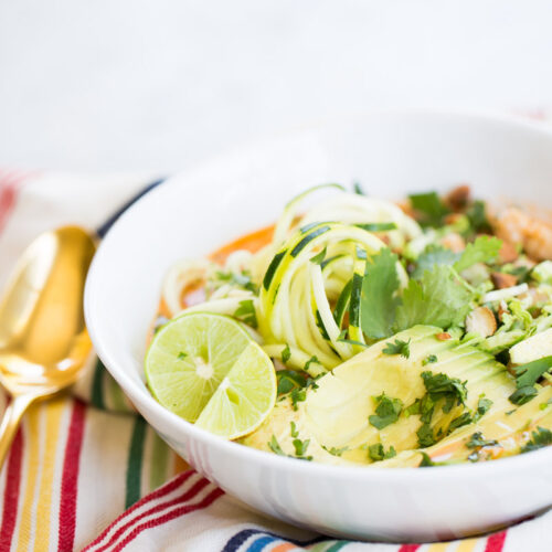 A white bowl filled with spiralized zucchini, avocado slices, lime wedges, fresh cilantro, and chopped nuts sits on a colorful striped napkin—an inspiring scene for anyone seeking a vibrant chia recipe beyond classic overnight oats.