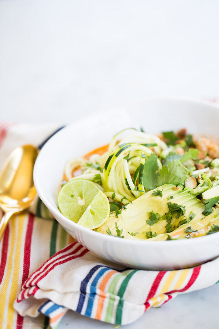 A white bowl filled with spiralized zucchini, avocado slices, lime wedges, fresh cilantro, and chopped nuts sits on a colorful striped napkin—an inspiring scene for anyone seeking a vibrant chia recipe beyond classic overnight oats.