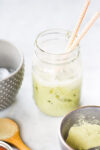 A mason jar filled with iced Matcha Latte and two patterned straws sits on a white surface. Nearby are a gray polka dot bowl with ice, a wooden spoon, and a small bowl containing green matcha powder—an easy recipe to try at home.