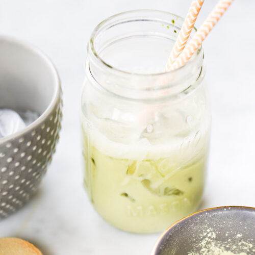 A mason jar filled with iced Matcha Latte and two patterned straws sits on a white surface. Nearby are a gray polka dot bowl with ice, a wooden spoon, and a small bowl containing green matcha powder—an easy recipe to try at home.