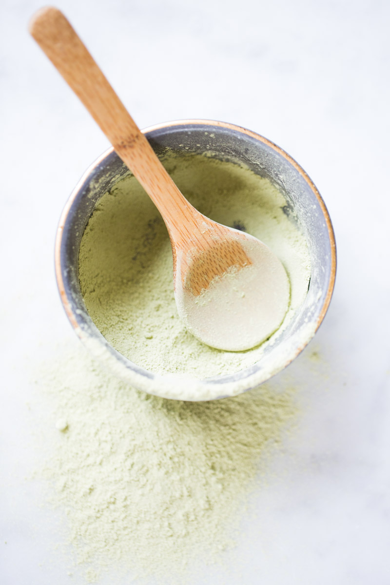 Un pequeño cuenco de metal lleno de polvo de matcha verde claro se encuentra sobre una superficie blanca, con algo de polvo derramado a su alrededor. Una cuchara de madera descansa dentro del cuenco, lista para recoger matcha fresco para su próximo vibrante matcha latte. La escena es luminosa y minimalista.
