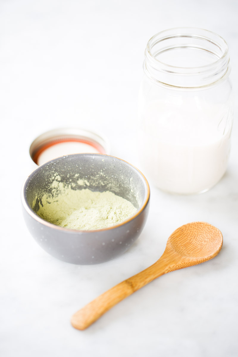 Un cuenco gris con polvo de matcha verde pálido, una cuchara de madera y un tarro abierto de leche están dispuestos sobre una superficie blanca: elementos esenciales perfectos para preparar un vibrante matcha latte en este entorno limpio y minimalista.