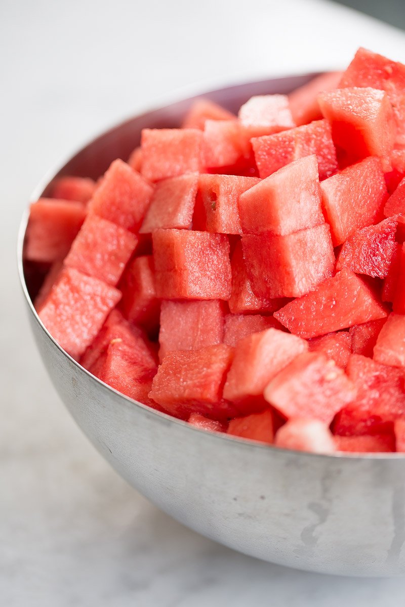 A stainless steel bowl brimming with evenly cut, vibrant red watermelon cubes, reminiscent of a refreshing watermelon poke. The juicy pieces display varying shades of red and pink, reflecting light beautifully. It rests on a slightly blurred surface, highlighting the freshness and texture.
