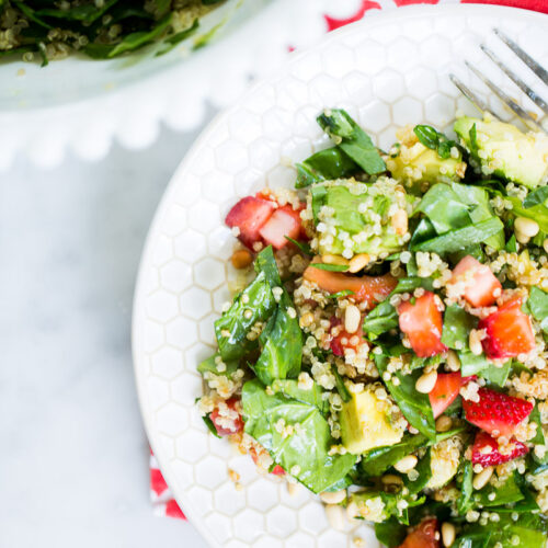 Primer plano de un plato blanco con ensalada de espinacas y fresas (quinoa, aguacate, espinacas, fresas picadas y piñones). El plato reposa sobre una servilleta roja estampada; un tenedor descansa en el borde y una ensaladera se asoma por una esquina.