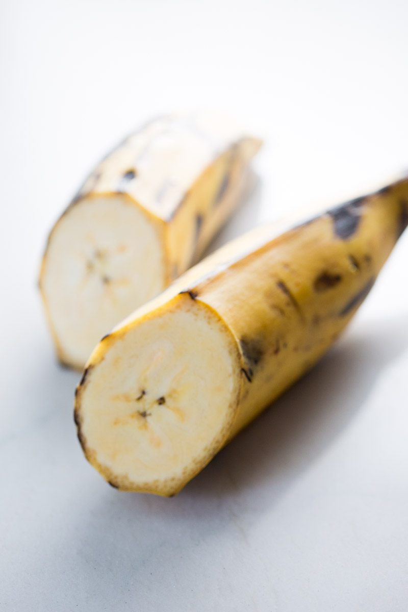 Close-up of a ripe plantain cut in half lengthwise on a white surface, much like the perfect accompaniment to enmoladas. The skin is yellow with brown spots, showing its ripeness. Its creamy interior boasts a soft texture, while the light background highlights the fruit's details.