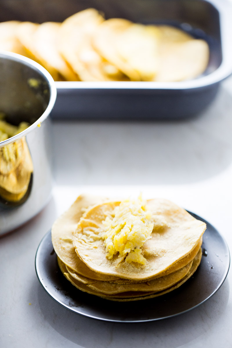 A stack of corn tortillas on a black plate with a scoop of mashed potatoes on top evokes the essence of enmoladas. In the background, a metal pot with more mashed potatoes and a baking dish with additional tortillas harmonize on the white countertop.