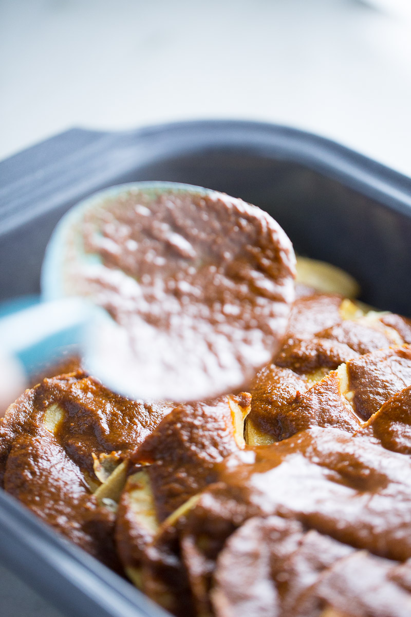 Close-up of a baking dish filled with sliced potatoes layered in a rich, reddish-brown mole sauce reminiscent of enmoladas. A spoon in the foreground drizzles more sauce over the top layer. The textured appearance of the potatoes and sauce promises they're perfectly ready for baking.