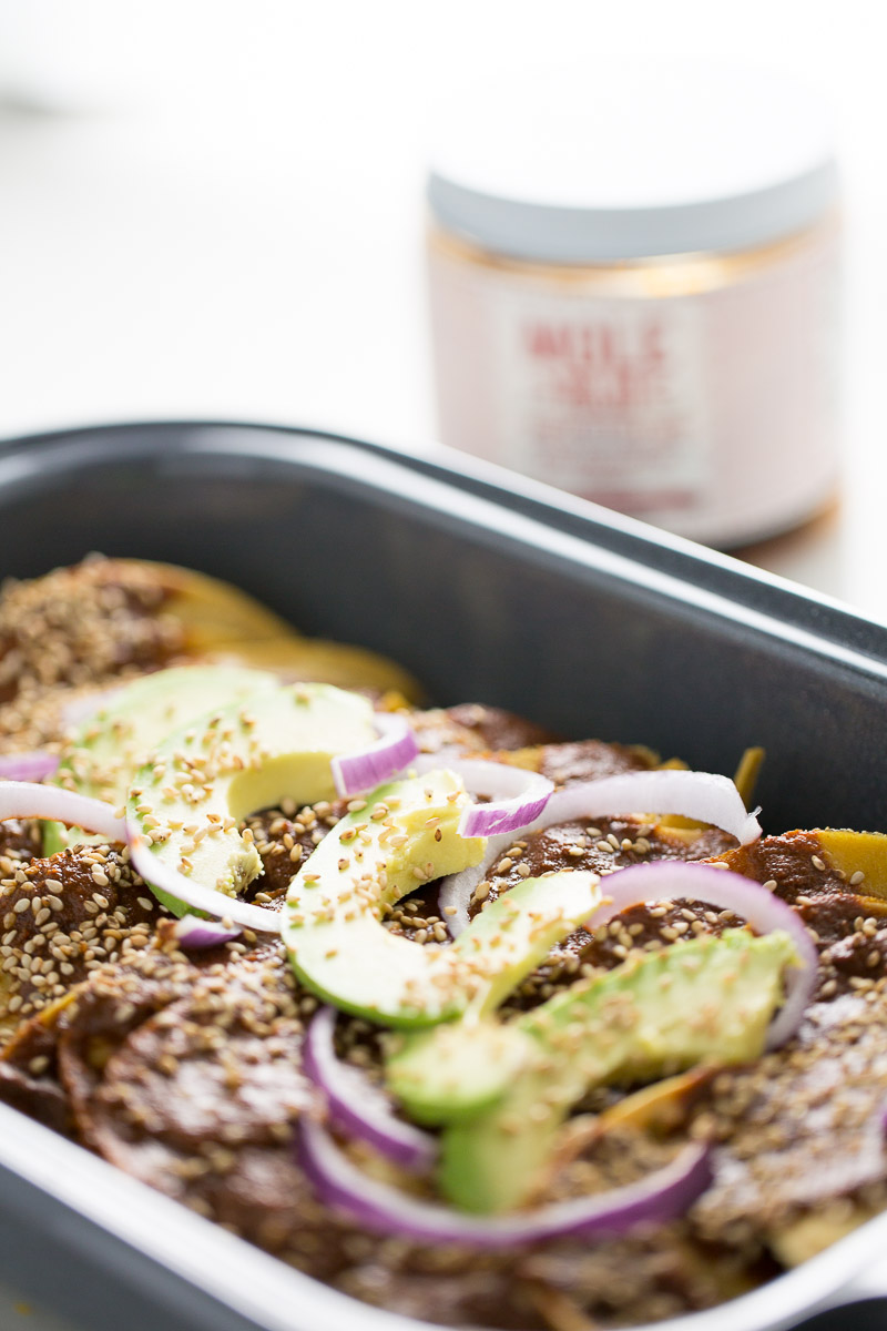 Close-up of enmoladas topped with slices of avocado, red onion, and sprinkled sesame seeds. The dish features a rich layer of mole sauce. In the blurred background, there's a jar with an unclear label. The focus is on the vibrant toppings and the dish's texture.