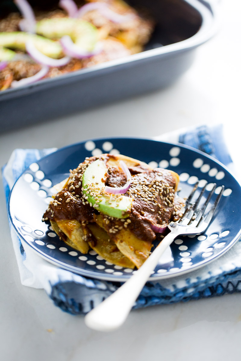 A blue plate with enmoladas, topped with rich mole sauce, sliced avocado, and red onion rings. The enchiladas are sprinkled with sesame seeds. A fork rests on the plate, which is set on a white table partially covered by a blue and white patterned cloth.