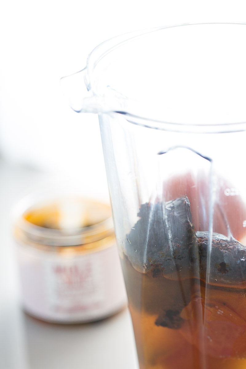 A clear blender container filled with dark, fibrous ingredients and liquid evokes the rich essence of mole enchiladas. In the background, a jar with a blurred label is slightly open, hinting at an enmoladas-related ingredient. The scene rests against a light backdrop, maintaining a clean, minimalist look.