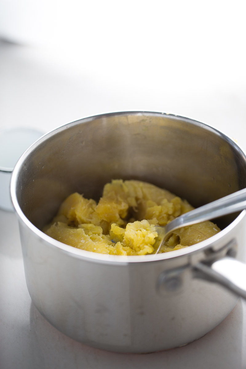 A silver pot on a white countertop contains mashed potatoes with a metal spoon, reminiscent of the comforting simplicity of enmoladas. The potatoes appear smooth with some chunks, indicating they have been partially mashed. A blurred jar lid is visible in the background, softly illuminated by gentle light.