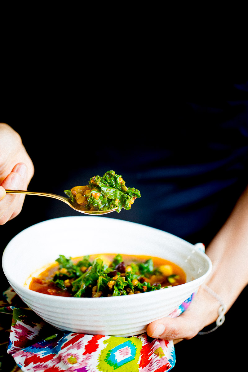 Una persona sostiene en una mano un cuenco estampado de sopa lentejas marroquí relleno de verduras de hoja verde, alubias y hortalizas, mientras levanta una cucharada de la sustanciosa y colorida mezcla con la otra, sobre un fondo negro liso.