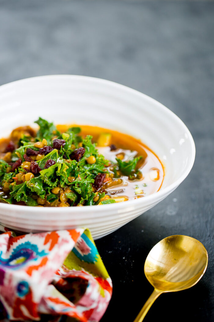 A white bowl filled with vibrant Moroccan Lentil Soup, topped with fresh kale, lentils, and dried cranberries sits on a dark surface. A patterned napkin and gold spoon complete the inviting vegan presentation.
