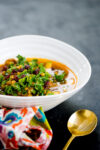 A white bowl filled with vibrant Moroccan Lentil Soup, topped with fresh kale, lentils, and dried cranberries sits on a dark surface. A patterned napkin and gold spoon complete the inviting vegan presentation.