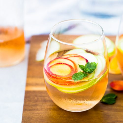 A stemless glass sits on a wooden board, filled with a light orange summer sangria. Garnished with thin slices of apple and peach, and topped with fresh mint leaves. Another drink and a clear glass container blur in the background.