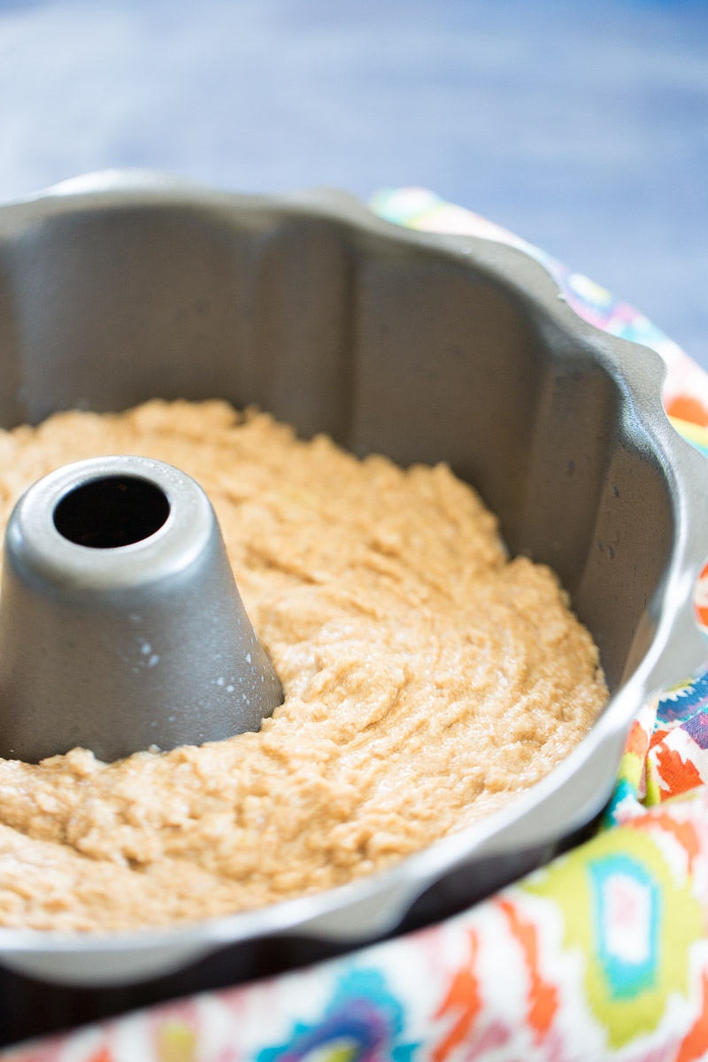 A gray Bundt cake pan filled with pale brown, textured healthy banana bread batter sits on a colorful patterned cloth. The batter is spread evenly around the pan’s central tube, ready to be baked. The background is softly blurred in shades of blue.