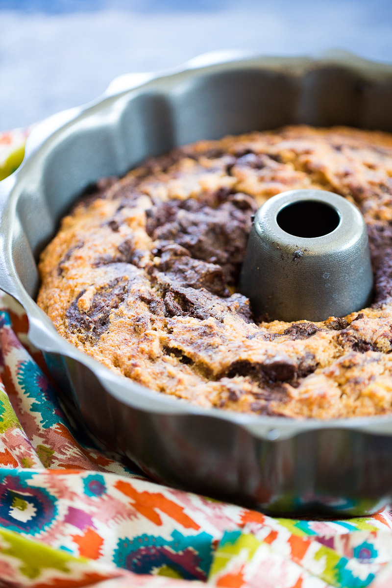 A close-up of marbled chocolate banana bread in a metal bundt pan, resting on a colorful, patterned cloth. The cake features swirled light and dark batter, a crispy golden-brown top, and the classic central hole from the pan.