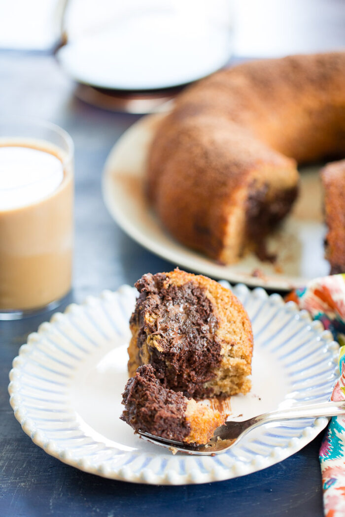 A fork holds a bite of marbled chocolate and vanilla bundt cake on a white plate. In the background, coffee with milk and the rest of the bundt cake, reminiscent of chocolate banana bread, sit near a colorful napkin.