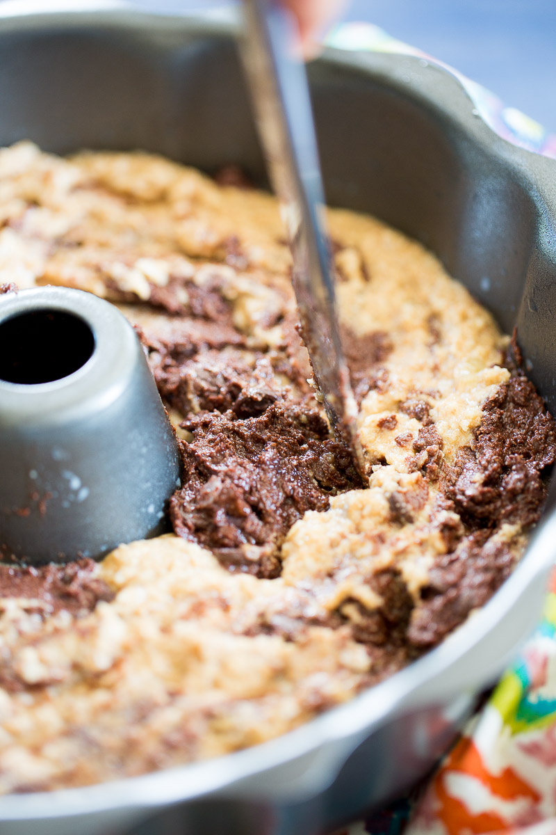 A close-up of Marbled Chocolate Banana Bread batter—light and chocolate—swirled in a Bundt pan. A hand swirls the mix before baking. The metallic gray pan features a central tube, with bright colors softly blurred in the corner.