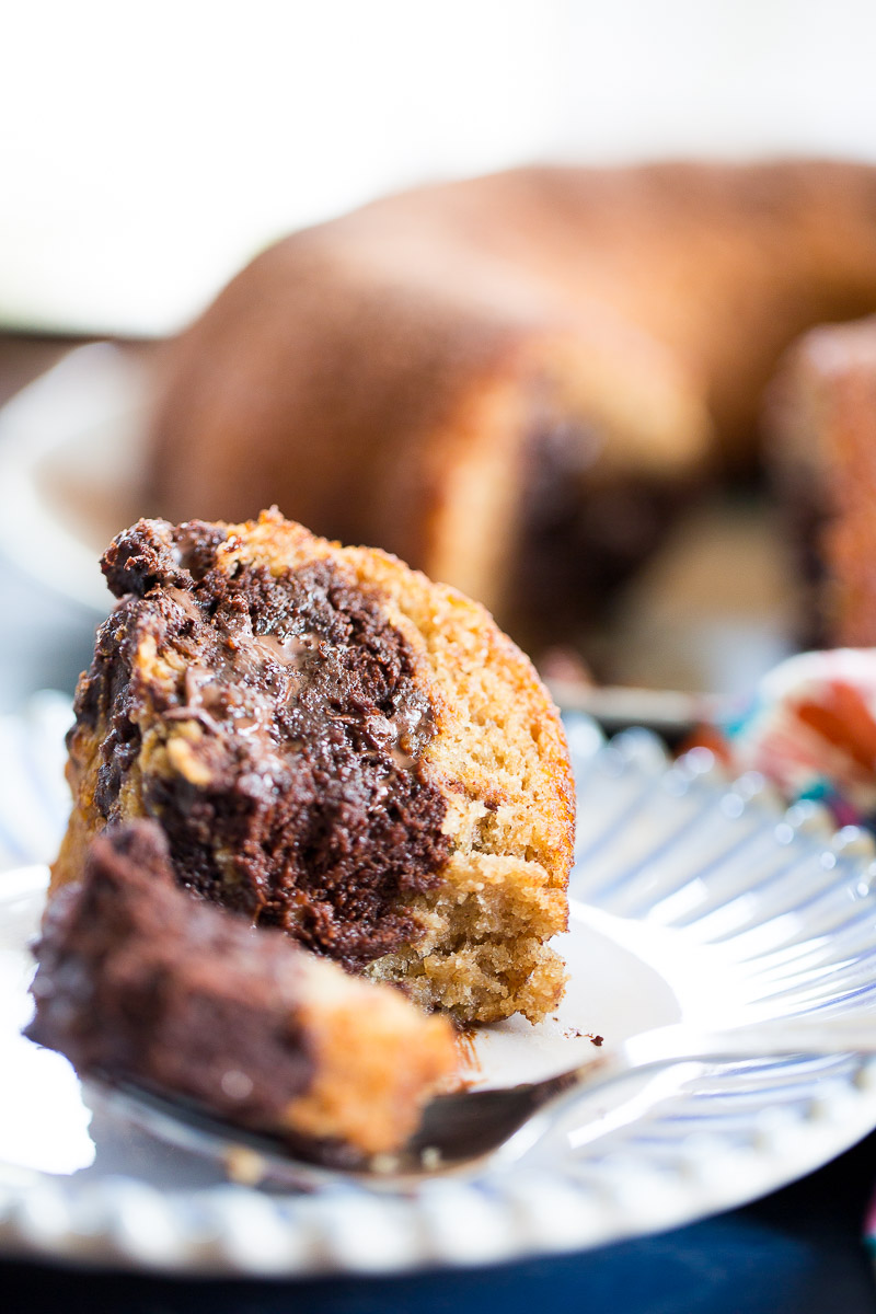 A close-up of a slice of chocolate banana bread bundt cake on a white plate, showing swirls of chocolate and yellow cake. A fork with a bite rests in front, with more cake on a platter in the blurred background. The texture appears moist and fluffy.