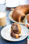 A fork holds a bite of marbled chocolate and vanilla bundt cake on a white plate. In the background, coffee with milk and the rest of the bundt cake, reminiscent of chocolate banana bread, sit near a colorful napkin.