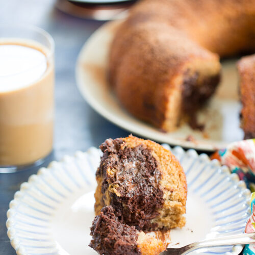 A fork holds a bite of marbled chocolate and vanilla bundt cake on a white plate. In the background, coffee with milk and the rest of the bundt cake, reminiscent of chocolate banana bread, sit near a colorful napkin.