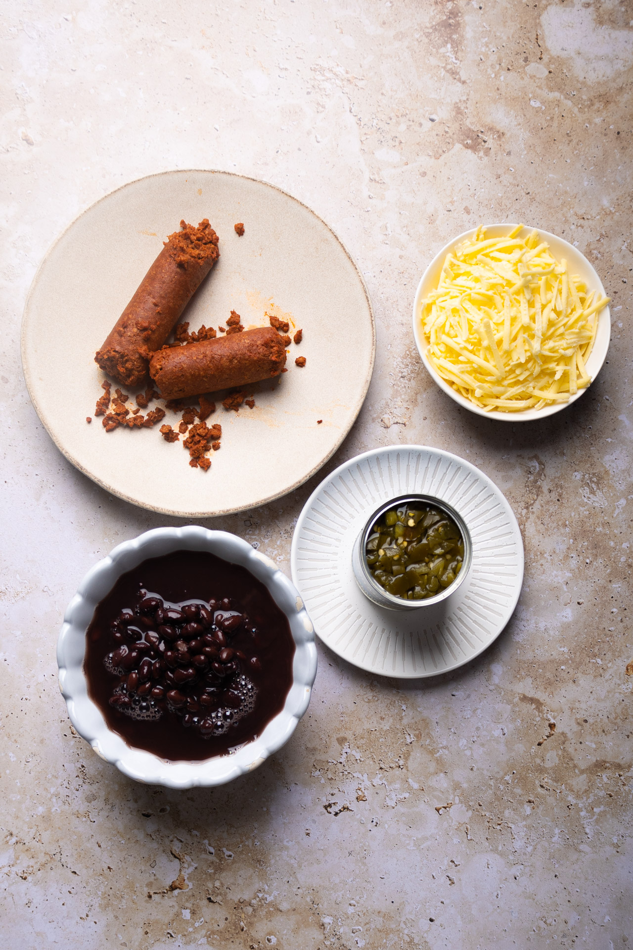 A flat lay of food ingredients on a textured surface features a plate with two cooked sausage links and crumbs, a bowl of shredded cheese, baked beans reminiscent of Mexican loaded beans, and a dish with chopped green chilies. The items are arranged in a square-like formation.