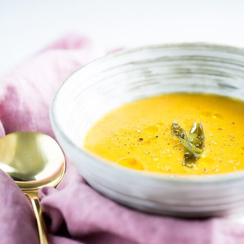 A textured white bowl filled with creamy roasted butternut squash soup (vegan), garnished with a sprig of sage and sprinkled with black pepper. The bowl rests on a soft pink cloth alongside a shiny gold spoon, all set against a bright, minimalist background.