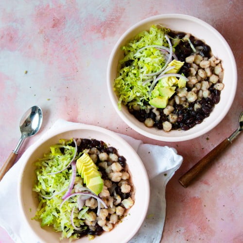 Two white bowls filled with black bean soup, white hominy, shredded lettuce, sliced avocado, and thin onion strips sit on a pink textured surface. Artfully arranged, the bowls evoke a fresh twist on classic pozole.
